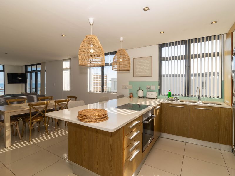 Modern open-plan kitchen and dining area featuring wood-finish cabinetry, stone countertops, integrated appliances, and large windows with vertical blinds.