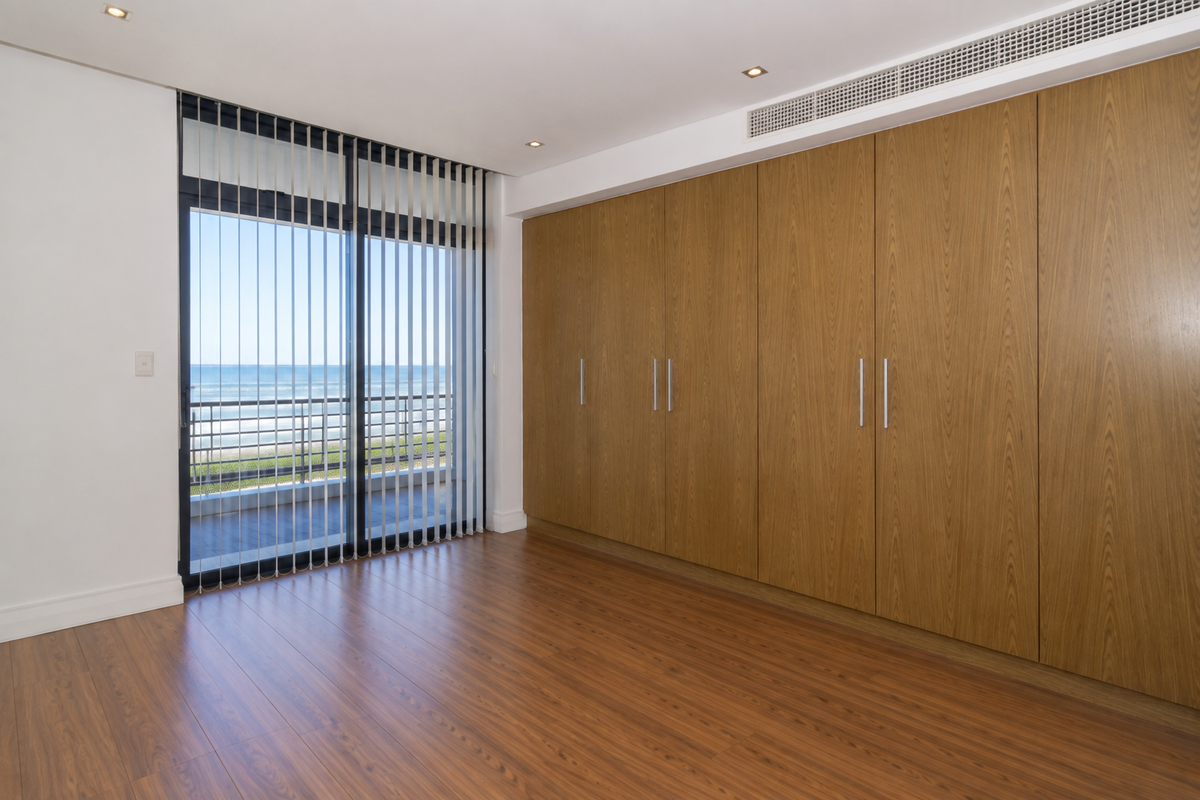 A bright, empty bedroom featuring modern wood-grain built-in wardrobes, laminate flooring, ducted air conditioning, and a sliding door leading to a balcony with ocean views.