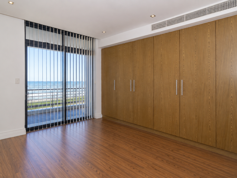 A bright, empty bedroom featuring modern wood-grain built-in wardrobes, laminate flooring, ducted air conditioning, and a sliding door leading to a balcony with ocean views.