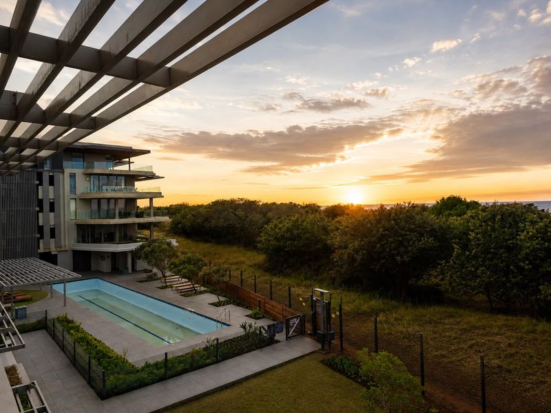 Elevated view of a modern apartment complex pool area featuring a lap pool, pergola, and landscaped grounds at sunset.