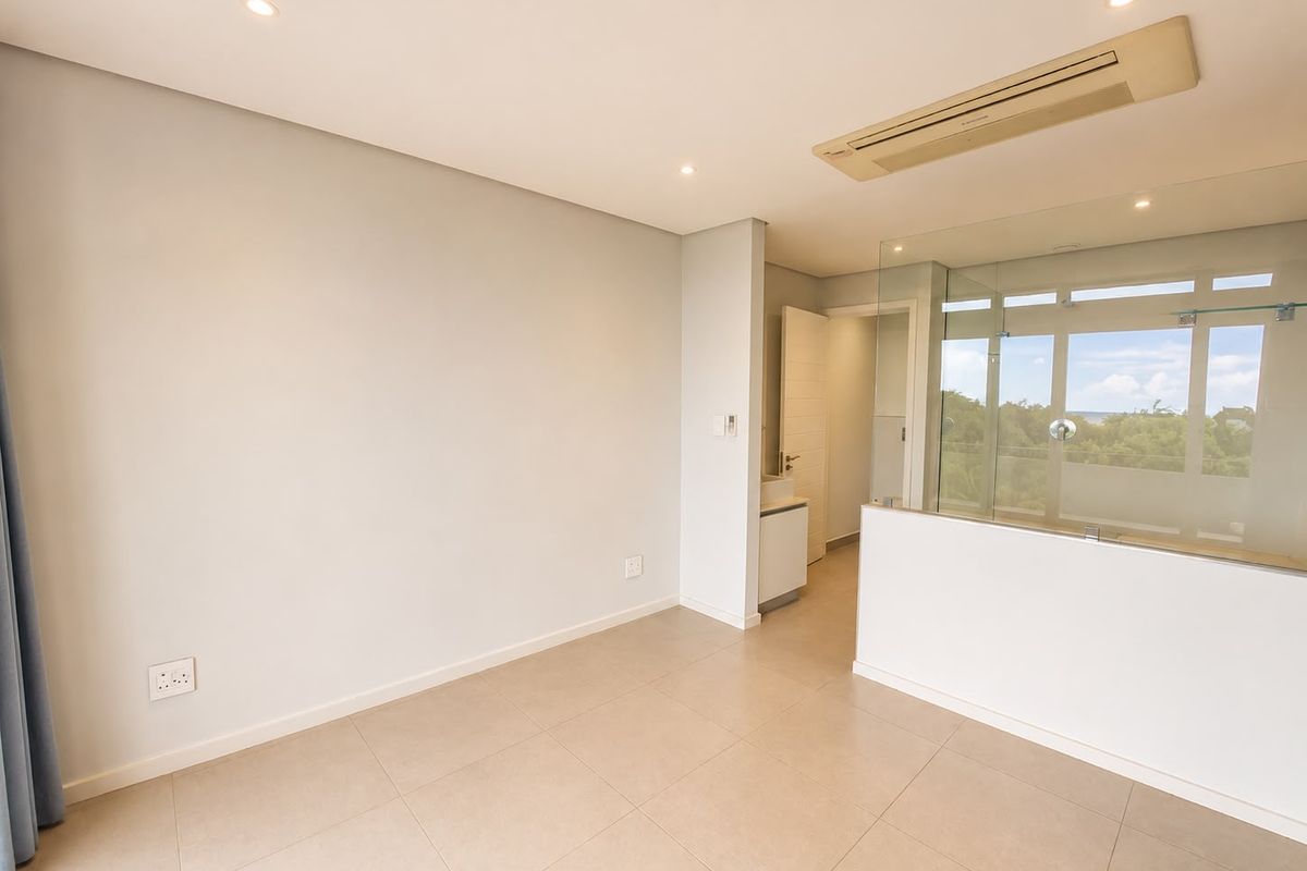 A bright, modern bedroom featuring neutral tiled flooring, recessed lighting, ducted air conditioning, and a glass-partitioned en-suite bathroom.
