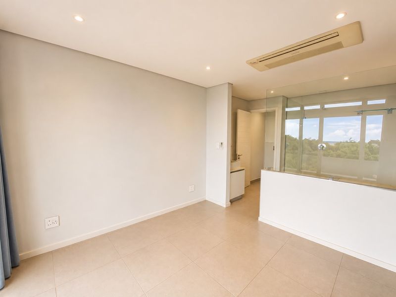 A bright, modern bedroom featuring neutral tiled flooring, recessed lighting, ducted air conditioning, and a glass-partitioned en-suite bathroom.