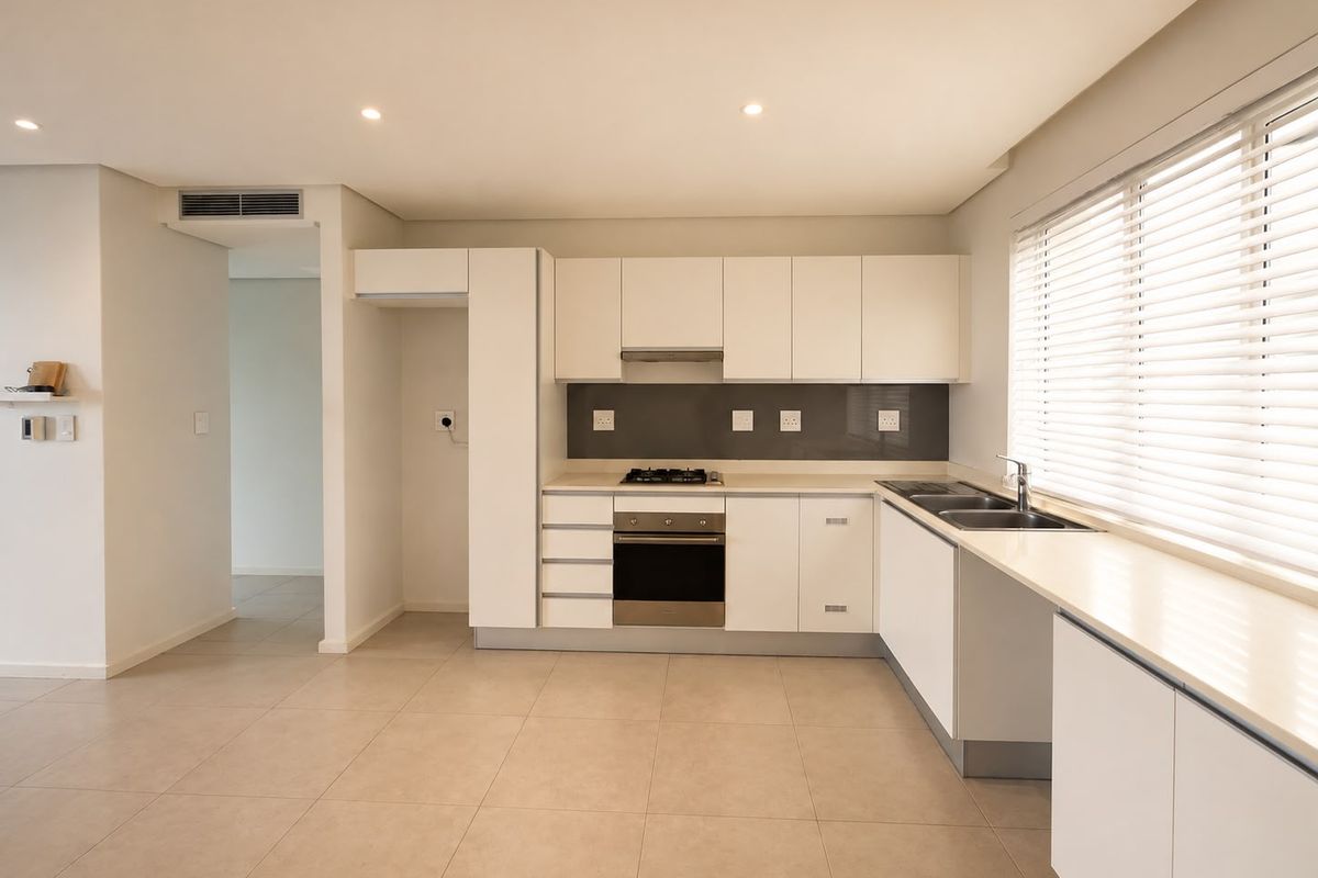 Modern, well-maintained kitchen featuring white cabinetry, stone countertops, built-in oven, gas hob, and a double sink with large windows and venetian blinds.