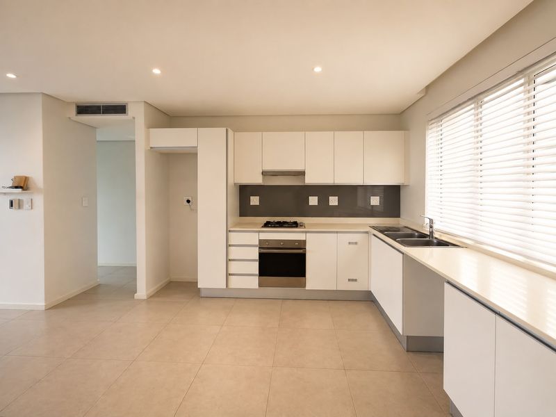 Modern, well-maintained kitchen featuring white cabinetry, stone countertops, built-in oven, gas hob, and a double sink with large windows and venetian blinds.