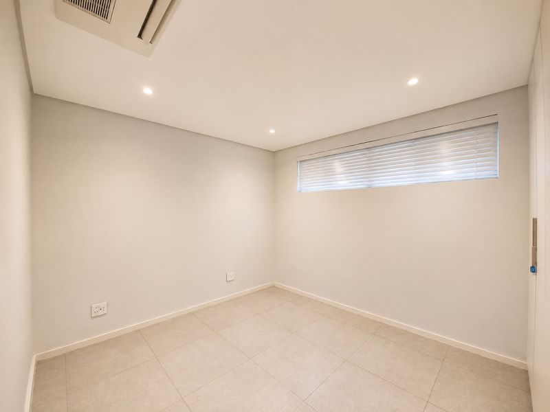 A clean, empty bedroom featuring neutral tiled flooring, recessed lighting, ducted air conditioning, and a built-in wardrobe with venetian blinds.