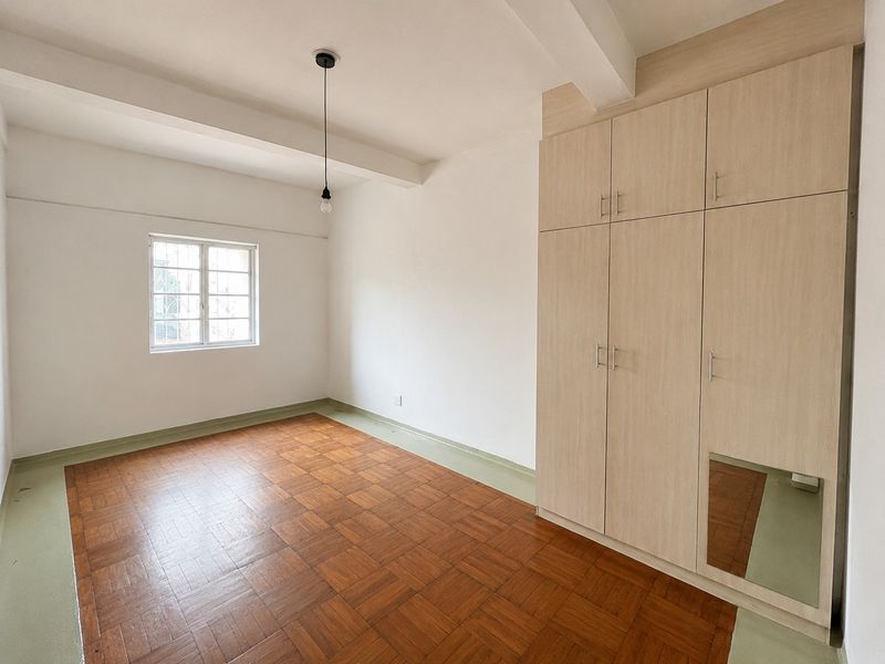A bright, empty bedroom featuring classic parquet flooring, a large built-in wardrobe with a mirror panel, and a window with security bars.