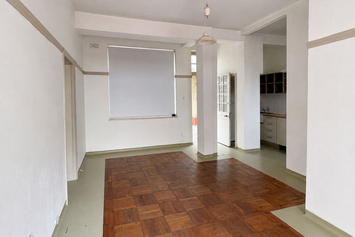An empty open-plan living area featuring original parquet flooring, neutral walls, and a view into the adjacent kitchen space.