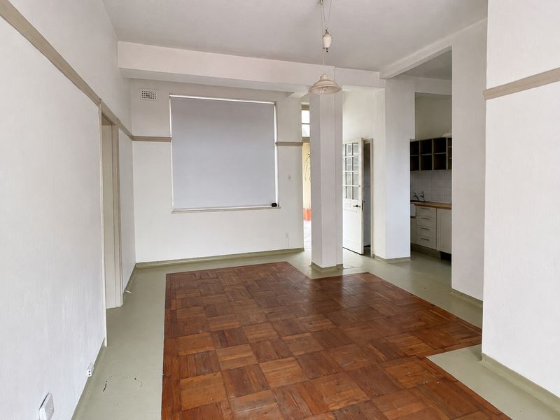 An empty open-plan living area featuring original parquet flooring, neutral walls, and a view into the adjacent kitchen space.