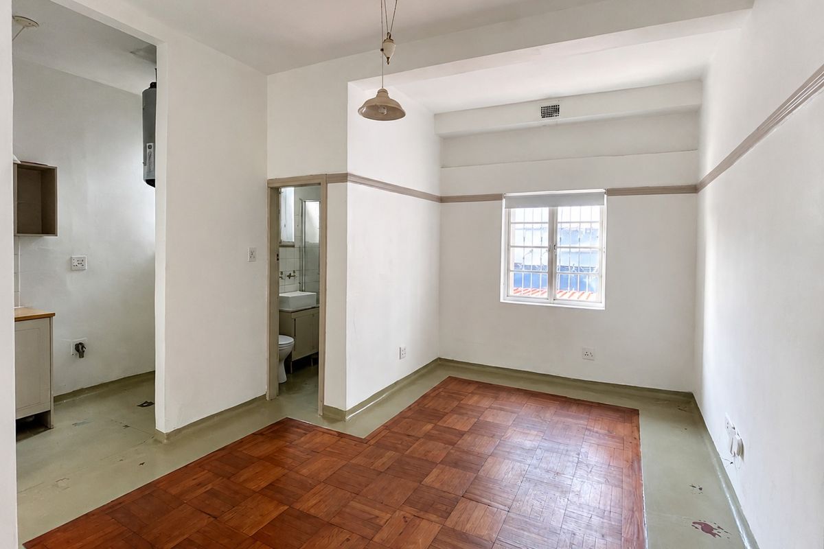 An empty open-plan living area featuring parquet flooring, neutral walls, and direct access to a small kitchen and bathroom.