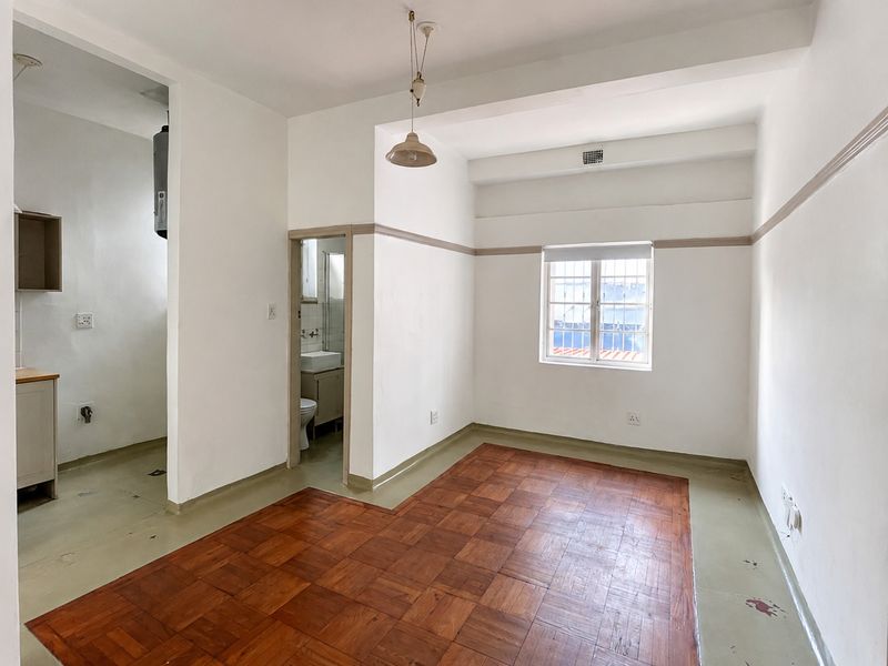 An empty open-plan living area featuring parquet flooring, neutral walls, and direct access to a small kitchen and bathroom.