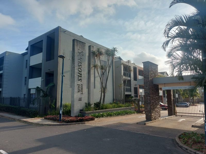 Front entrance view of Seasons Court apartment complex, featuring a secure gated access point, stone-clad pillars, and modern concrete building facade.