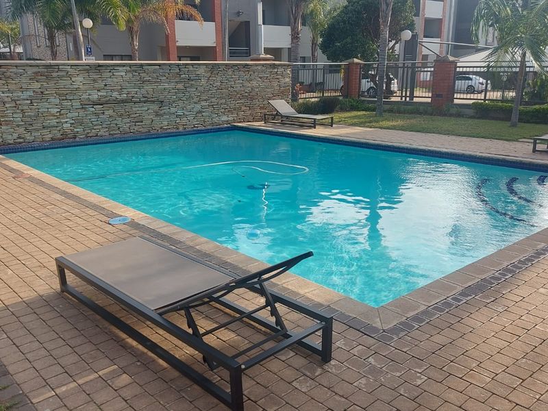 Communal swimming pool area featuring a paved deck, stone-clad feature wall, and landscaped surroundings within the apartment complex.