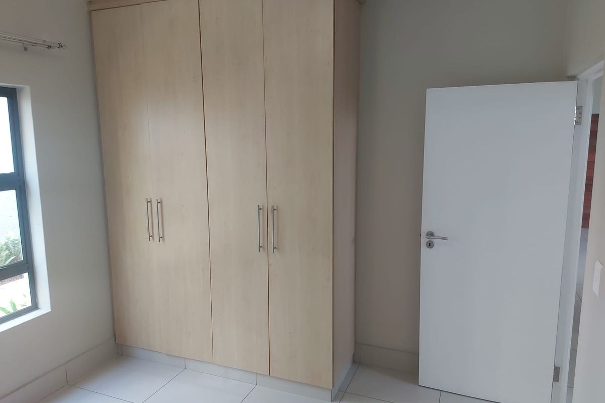 A clean, empty bedroom featuring light wood-finish built-in cupboards, neutral tiled flooring, and a modern aluminium window frame.