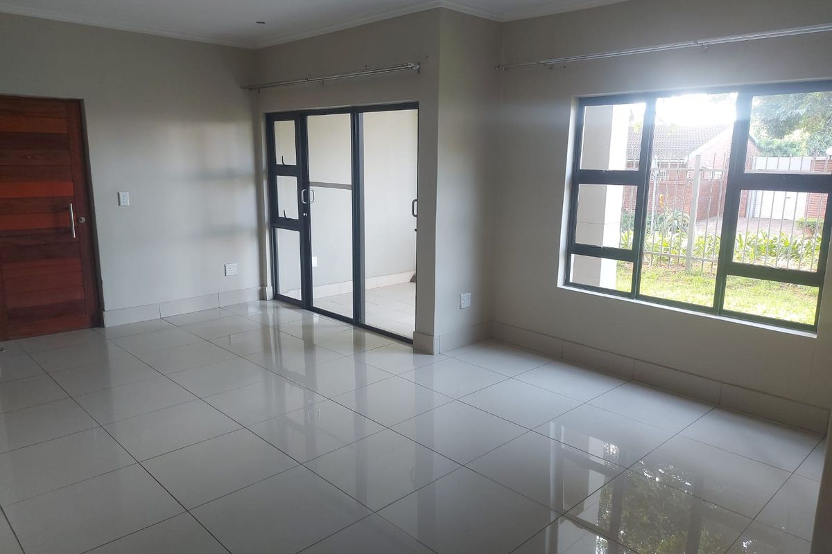 A bright, empty living area featuring neutral tiled flooring, modern aluminium window frames, and a sliding door leading to an outdoor space.