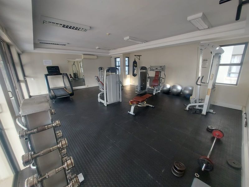 A functional apartment gym facility featuring rubber flooring, wall-mounted mirrors, and integrated air conditioning, equipped with various fitness machines and weights.