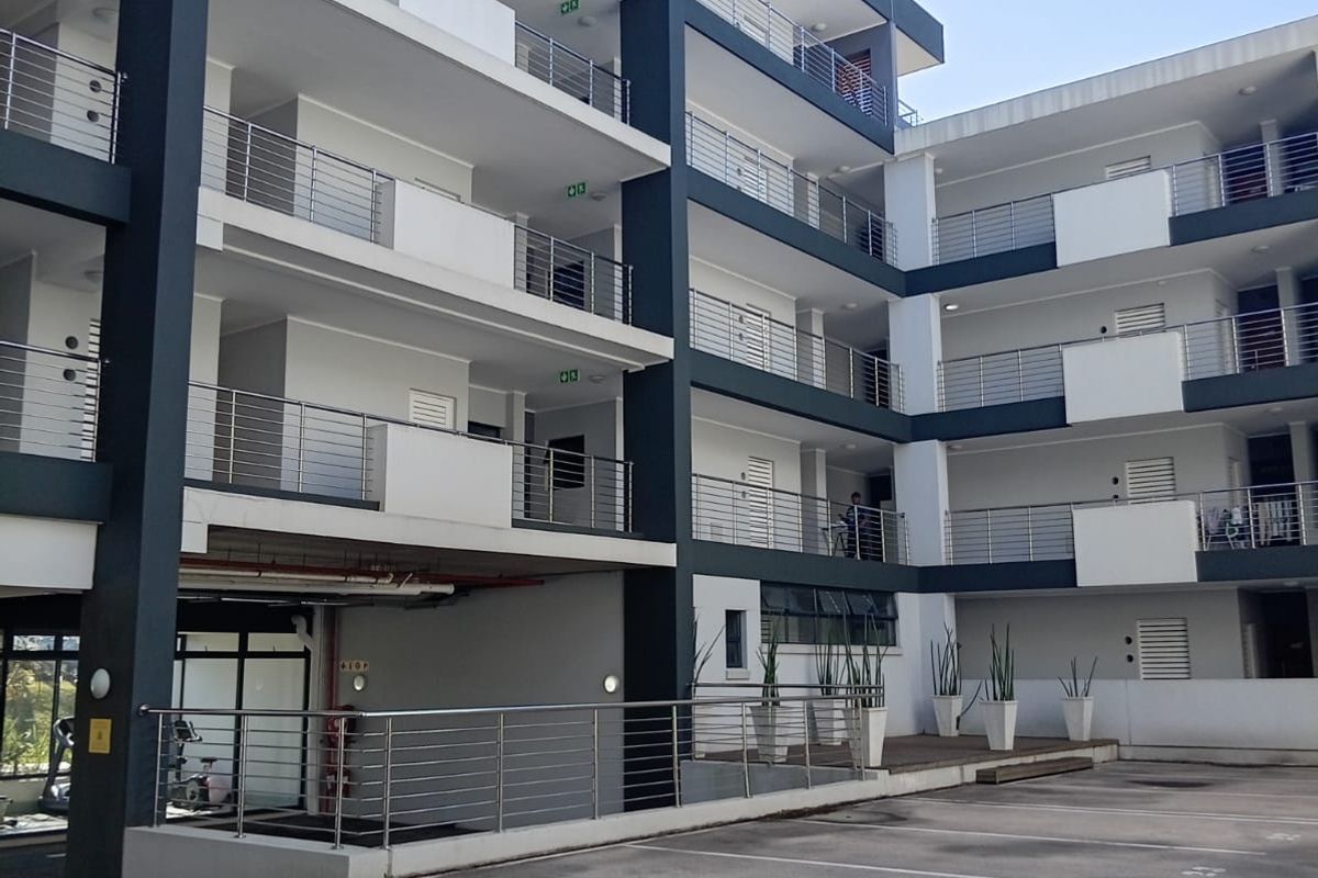 Exterior view of a modern apartment complex featuring multi-level balconies with stainless steel railings and a paved ground-level parking area.