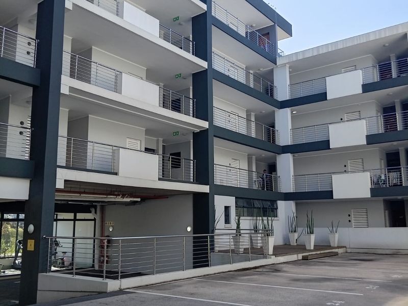 Exterior view of a modern apartment complex featuring multi-level balconies with stainless steel railings and a paved ground-level parking area.
