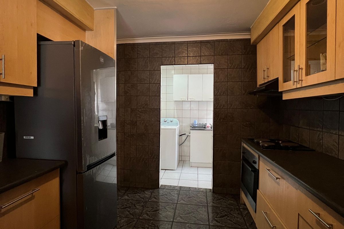 A functional galley-style kitchen featuring wood-finish cabinetry, dark tiled surfaces, and a built-in oven, with an adjoining scullery area visible through the doorway.