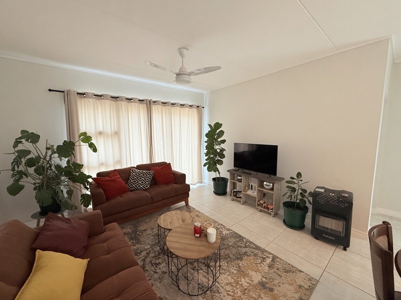 A bright and spacious living room featuring neutral tiled flooring, a ceiling fan, and large windows dressed with curtains.