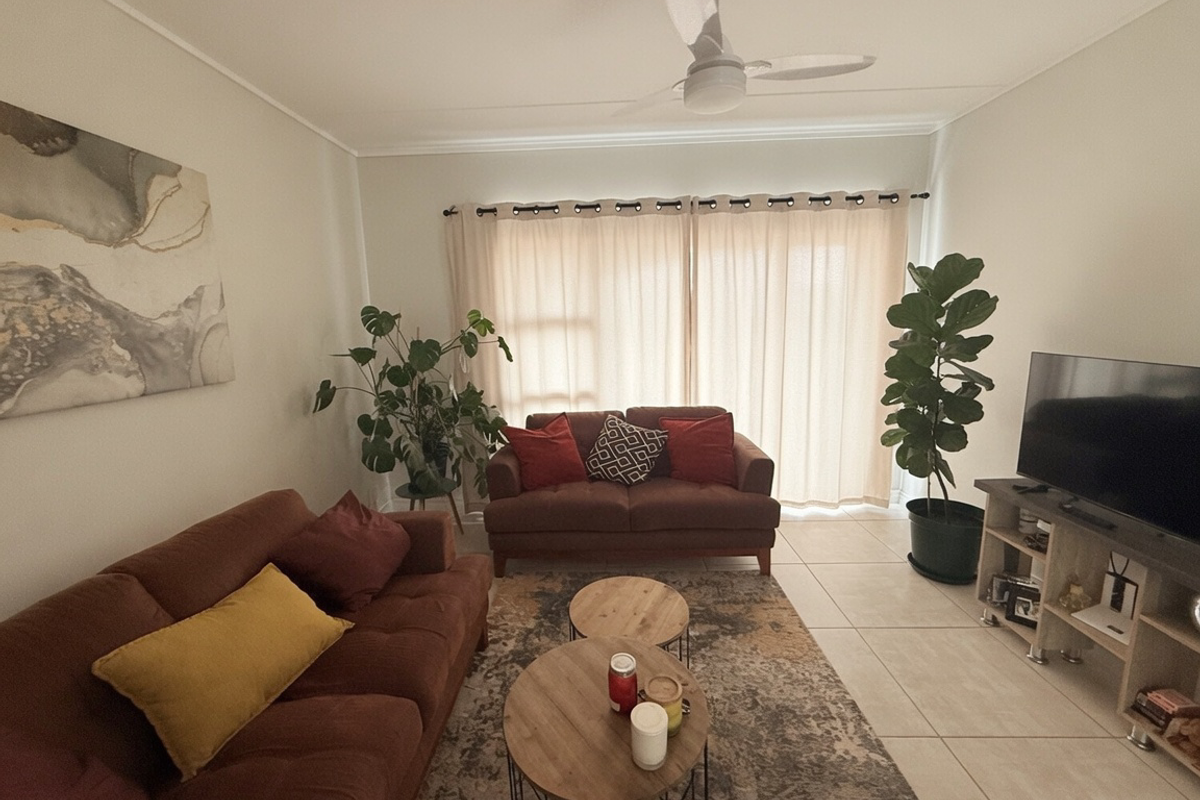 A comfortable living room featuring neutral tiled flooring, a ceiling fan, and large windows dressed with curtains, providing a bright and inviting space.