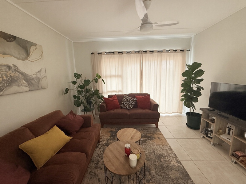 A comfortable living room featuring neutral tiled flooring, a ceiling fan, and large windows dressed with curtains, providing a bright and inviting space.