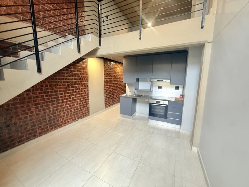Modern open-plan apartment featuring a sleek kitchen with built-in appliances, exposed brick feature wall, and a staircase leading to an upper level.