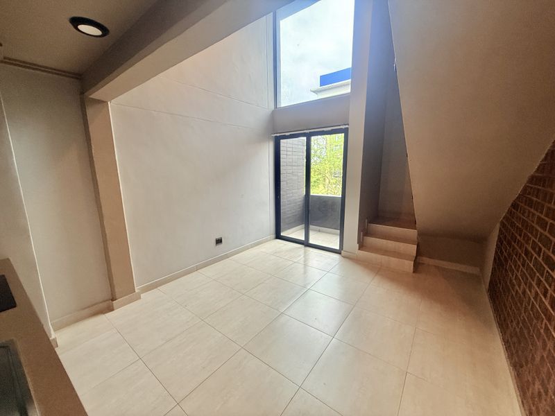 Spacious open-plan living area featuring double-volume ceilings, large windows for natural light, tiled flooring, and an exposed brick feature wall.