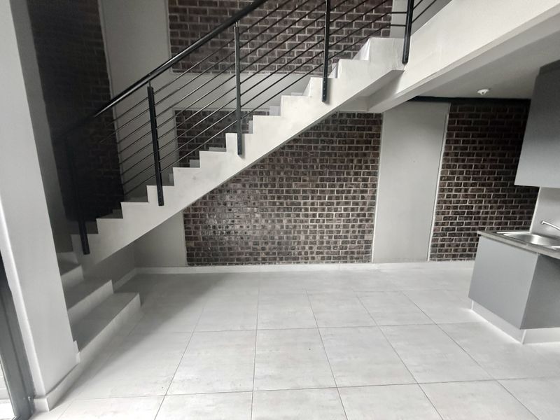 Open plan living area featuring a modern staircase with metal balustrade, exposed brick feature wall, tiled flooring, and a compact kitchen area.