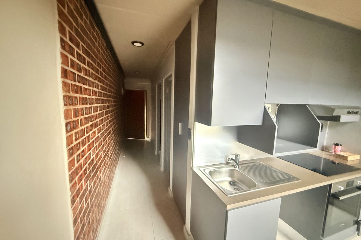 Compact kitchen area featuring modern grey cabinetry, integrated appliances, and a distinctive exposed brick feature wall leading into a hallway.