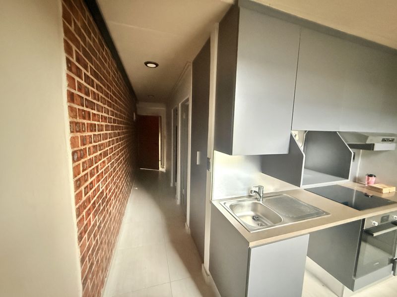 Compact kitchen area featuring modern grey cabinetry, integrated appliances, and a distinctive exposed brick feature wall leading into a hallway.
