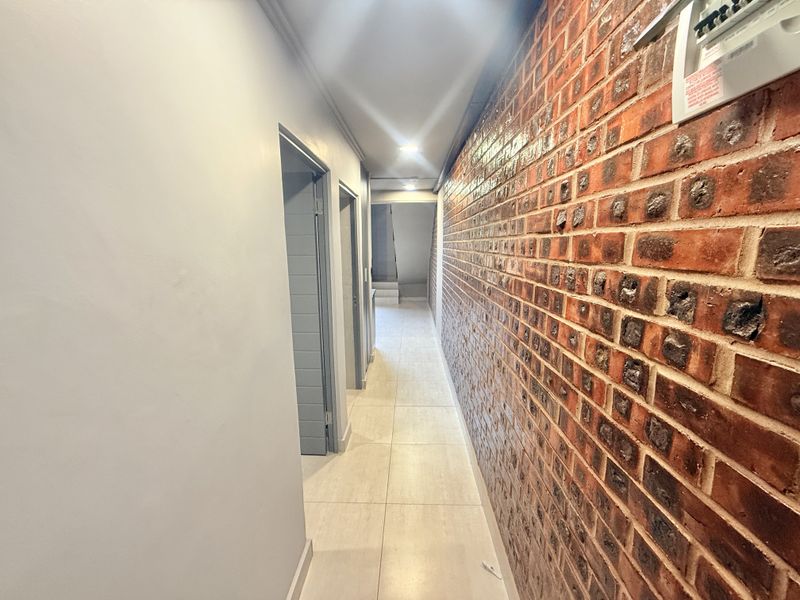 A clean, modern hallway featuring a striking exposed brick feature wall, neutral tiled flooring, and recessed ceiling lighting.