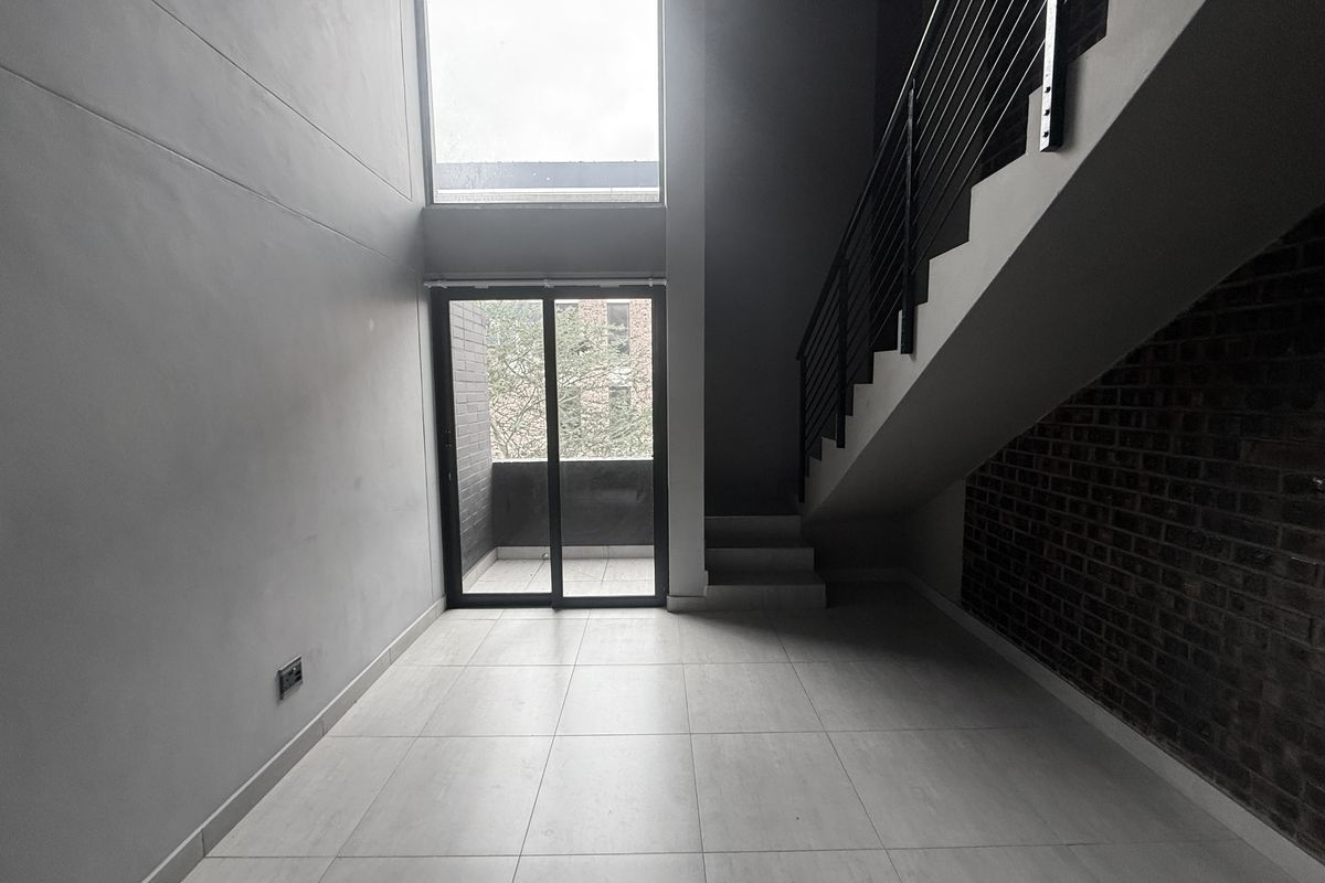 Modern double-volume living area featuring an exposed brick wall, sleek staircase with metal balustrade, large windows, and direct access to a balcony.