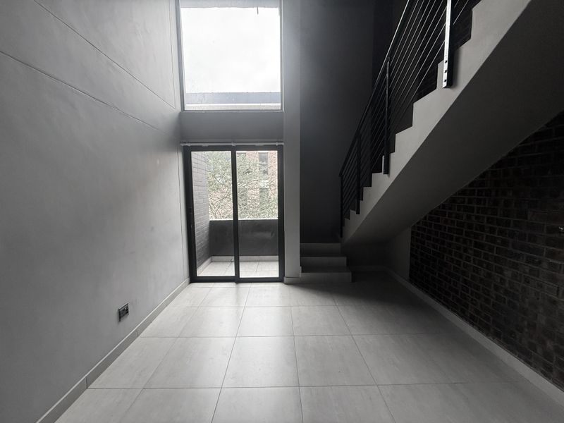 Modern double-volume living area featuring an exposed brick wall, sleek staircase with metal balustrade, large windows, and direct access to a balcony.