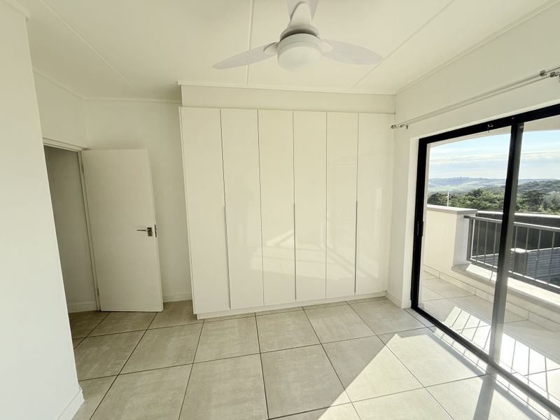 A bright, modern bedroom featuring built-in cupboards, a ceiling fan, tiled flooring, and a sliding glass door leading to a private balcony with scenic views.