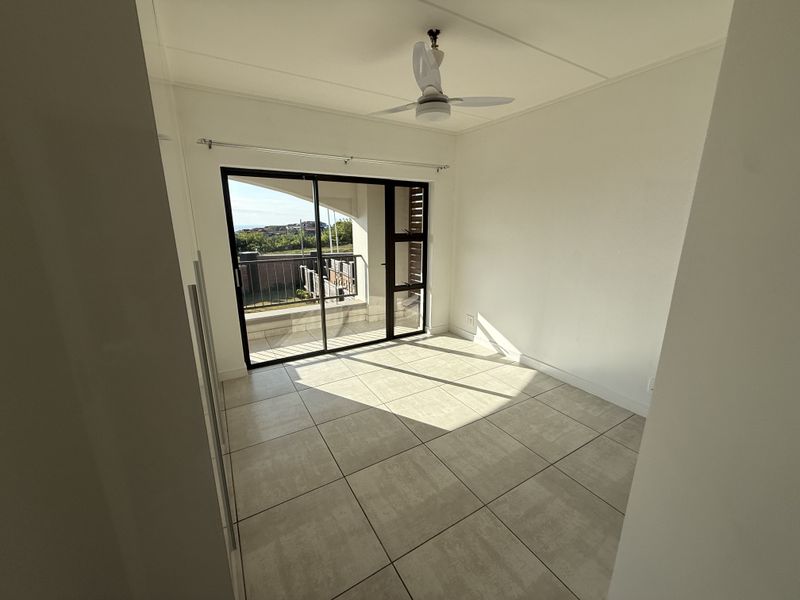 A bright, empty bedroom featuring neutral tiled flooring, a ceiling fan, and a sliding glass door providing access to an outdoor balcony.