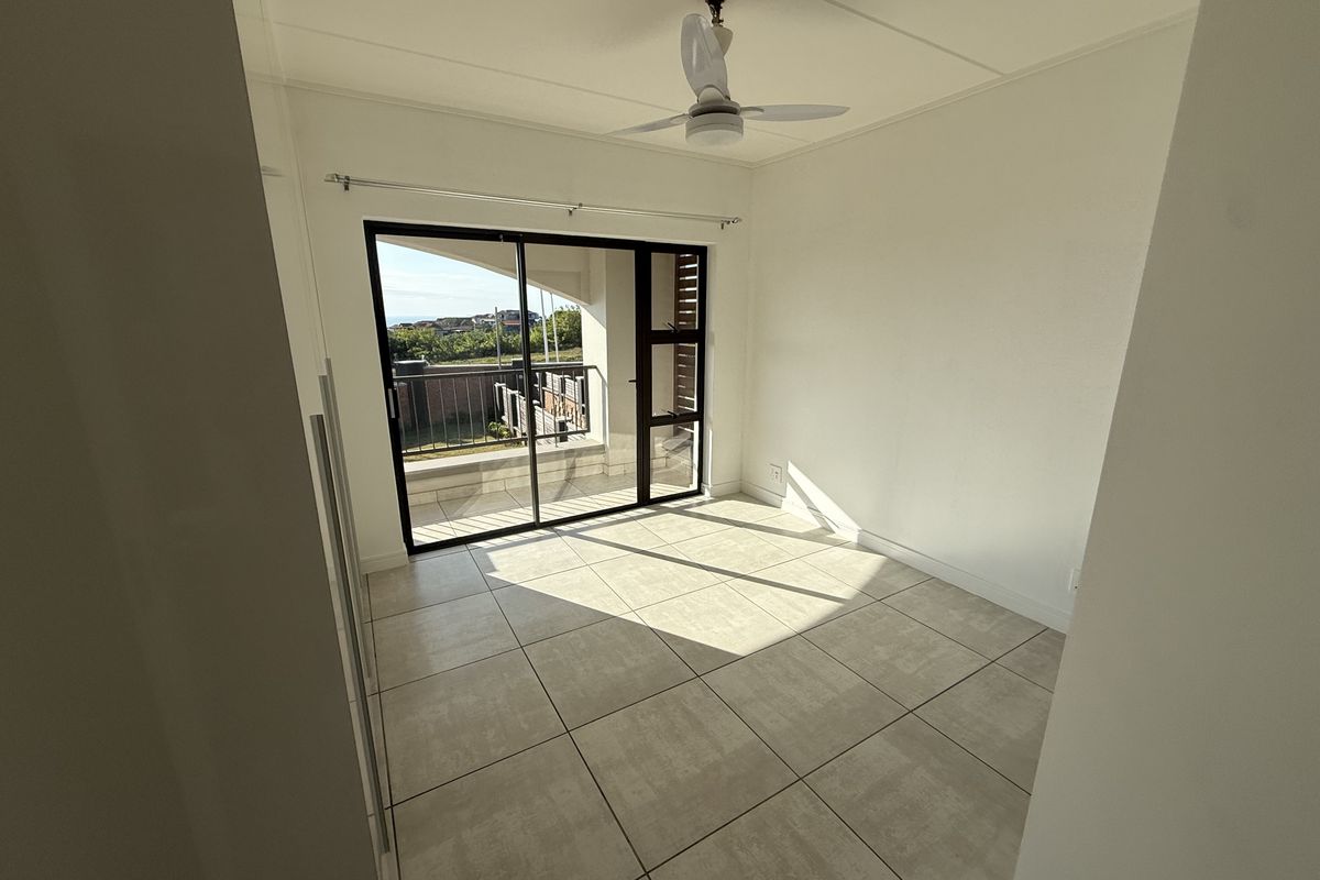 A bright, empty bedroom featuring neutral tiled flooring, a ceiling fan, and a sliding glass door providing access to an outdoor balcony.