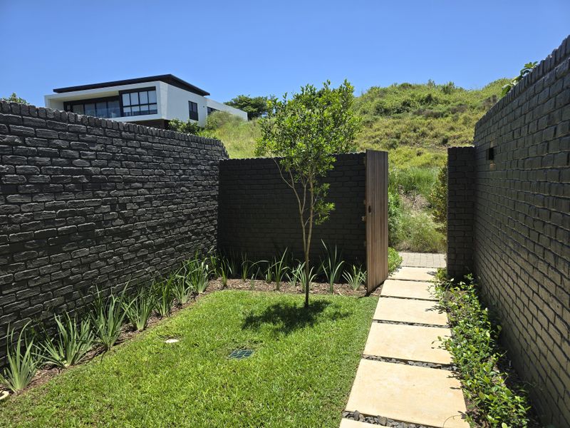A well-maintained private garden area featuring dark brick boundary walls, a stone paver pathway, and a wooden gate leading to the surrounding landscape.