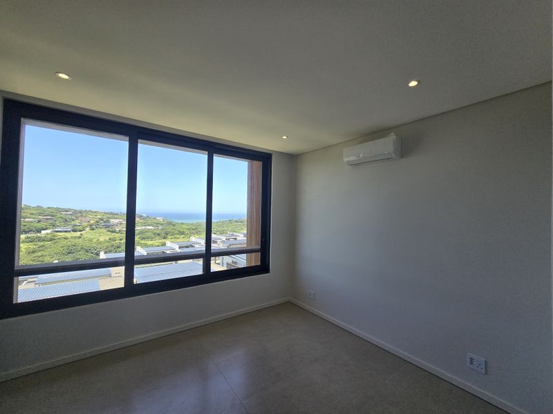 A bright, empty bedroom featuring tiled floors, recessed lighting, a wall-mounted air conditioning unit, and large windows offering scenic coastal views.