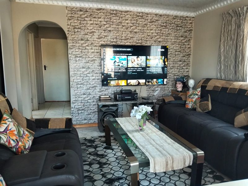 A living room featuring a brick-effect accent wall, decorative ceiling cornice, and tiled flooring, connected to an adjacent hallway via an arched doorway.