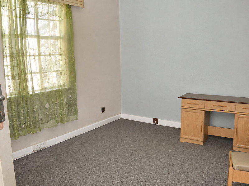 A sparsely furnished bedroom featuring carpeted flooring, a window with curtains, and basic electrical fixtures, showing signs of wear on the door frame.