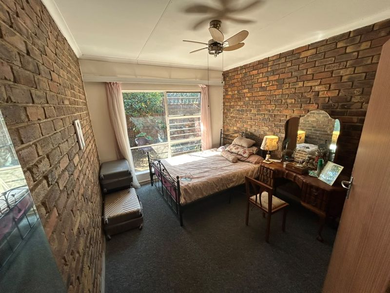 A bedroom featuring exposed brick walls, a ceiling fan, and large windows providing natural light and a view of the garden area.