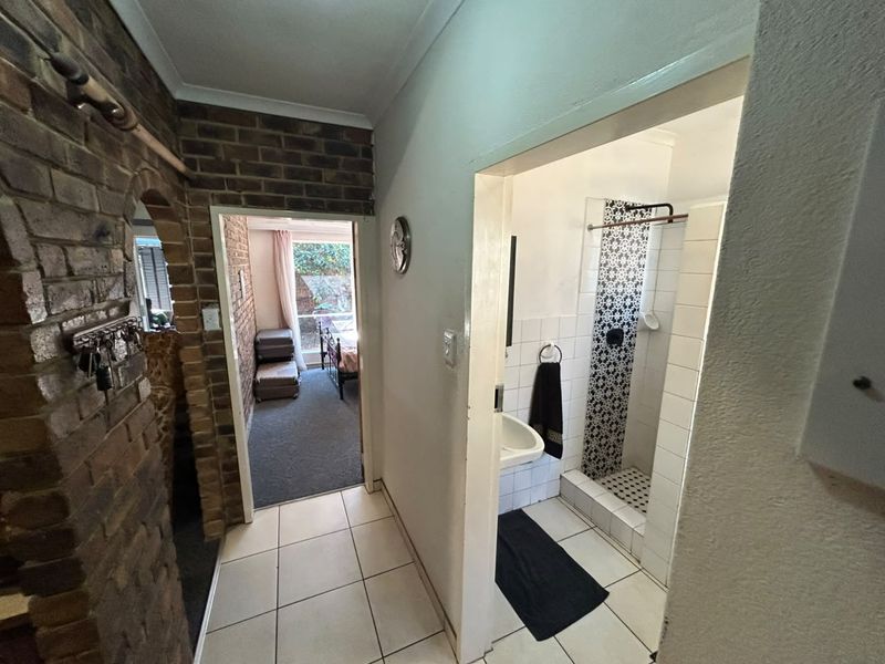 A hallway featuring exposed brickwork and tiled flooring, providing access to a bathroom with a shower and a bedroom beyond.