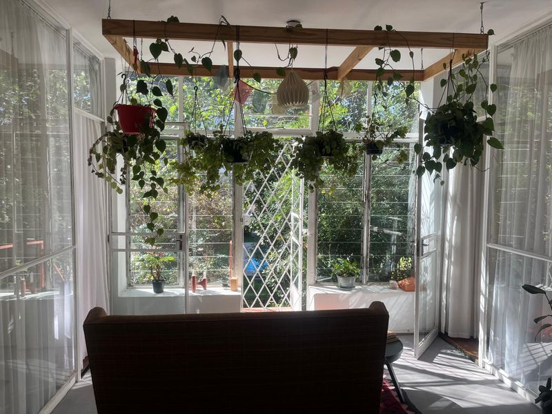 A sun-drenched enclosed patio or sunroom featuring large glass louvre windows, a timber overhead frame, and direct access to the garden.