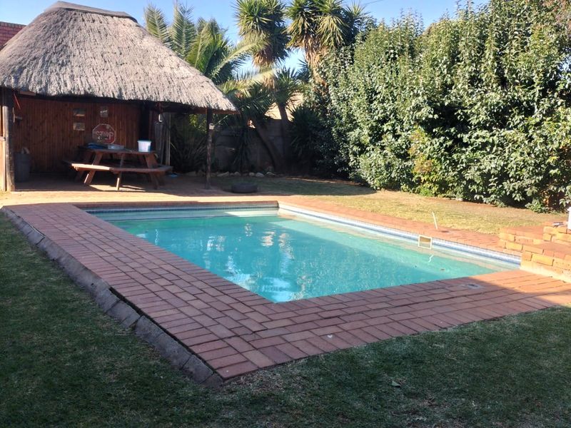 Outdoor pool area featuring a rectangular swimming pool with brick paving and an adjacent thatched lapa structure in a garden setting.