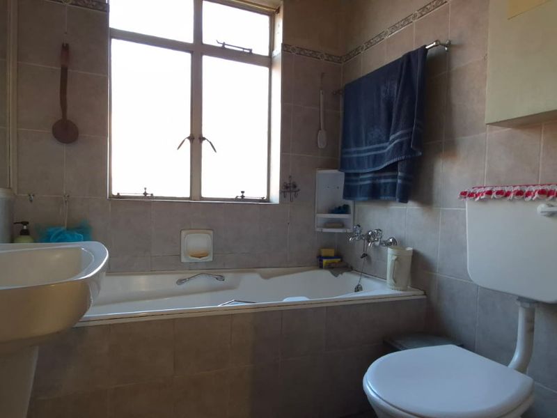 A dated bathroom featuring a pedestal basin, built-in bathtub, and a traditional toilet with a high-level cistern, finished with neutral wall tiles.