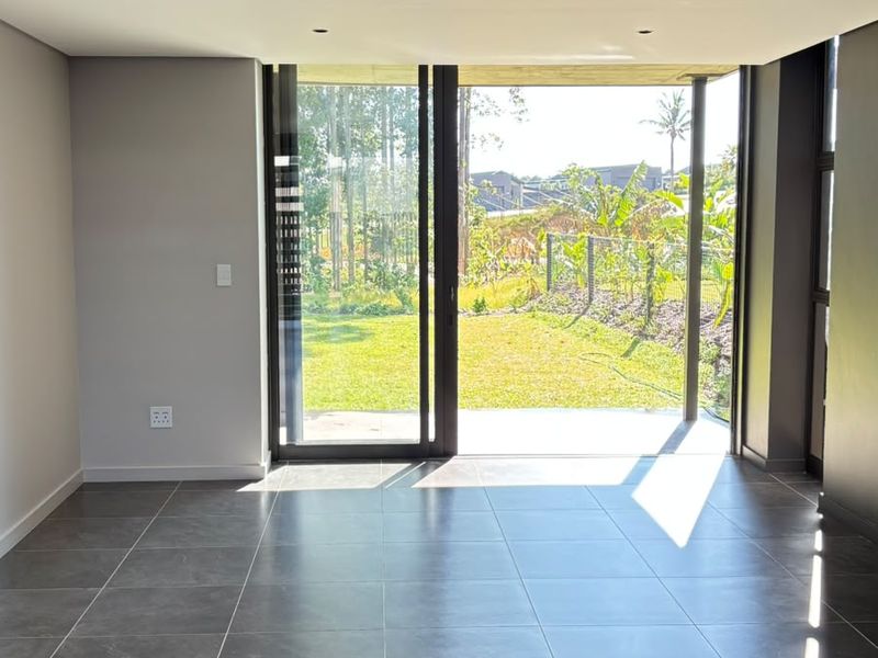 A bright, modern, and empty living area featuring large format floor tiles, recessed lighting, and expansive sliding glass doors opening to a garden view.