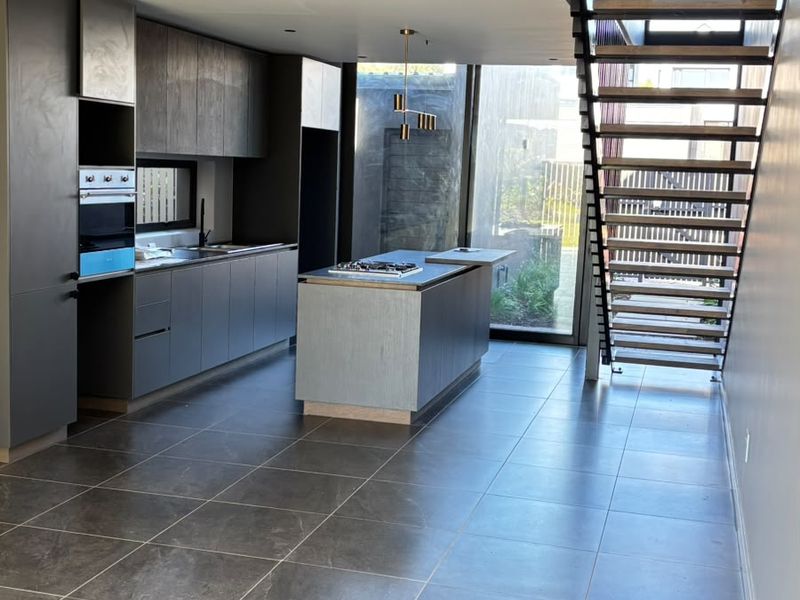 Modern open-plan kitchen featuring sleek dark cabinetry, a central island with cooktop, tiled flooring, and an adjacent architectural staircase.
