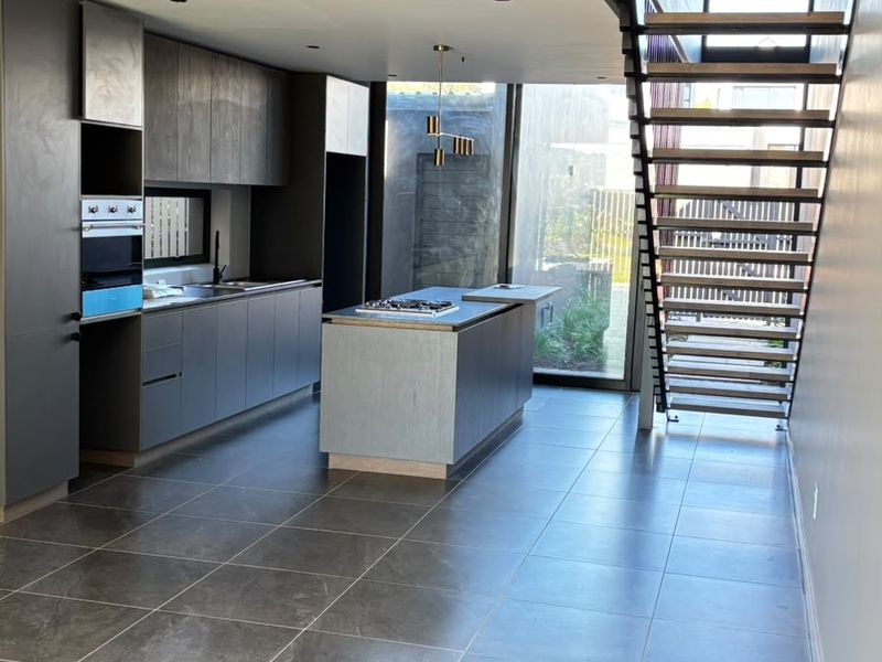 Modern open-plan kitchen featuring dark cabinetry, a central island with hob, and a striking floating staircase, finished with large-format floor tiles.