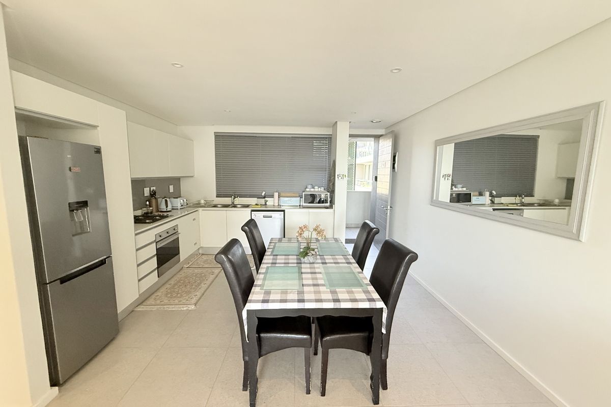 Modern open-plan kitchen and dining area featuring sleek white cabinetry, integrated appliances, tiled flooring, and recessed lighting, creating a bright and functional living space.