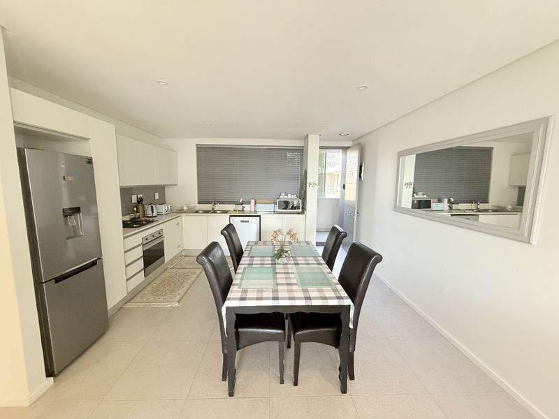 Modern open-plan kitchen and dining area featuring sleek white cabinetry, integrated appliances, tiled flooring, and recessed lighting, creating a bright and functional living space.